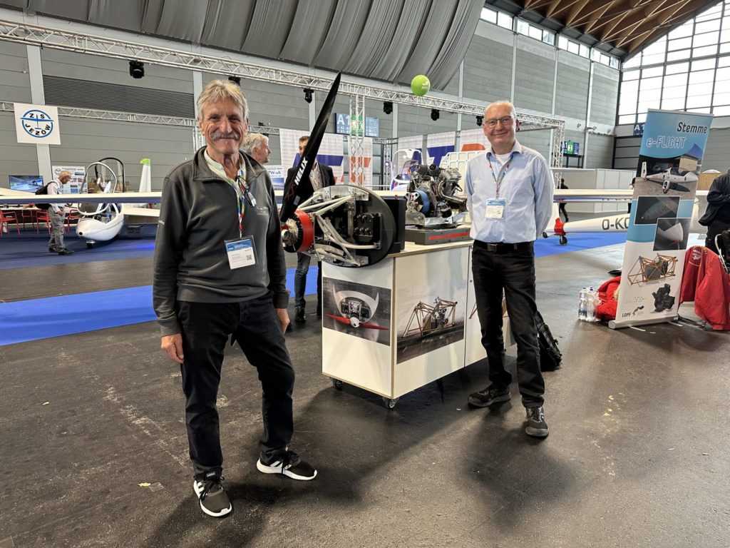 Klaus Ohlmann (links) und Karl Pickan (rechts) präsentieren vor einem Modell des innovativen Elektroantriebs, bestehend aus Batterien und einem Range Extender. Dieses Konzept verkörpert die Zukunft des emissionsarmen Segelfliegens, kombiniert traditionelle Glidertechnologie mit moderner elektrischer Effizienz.